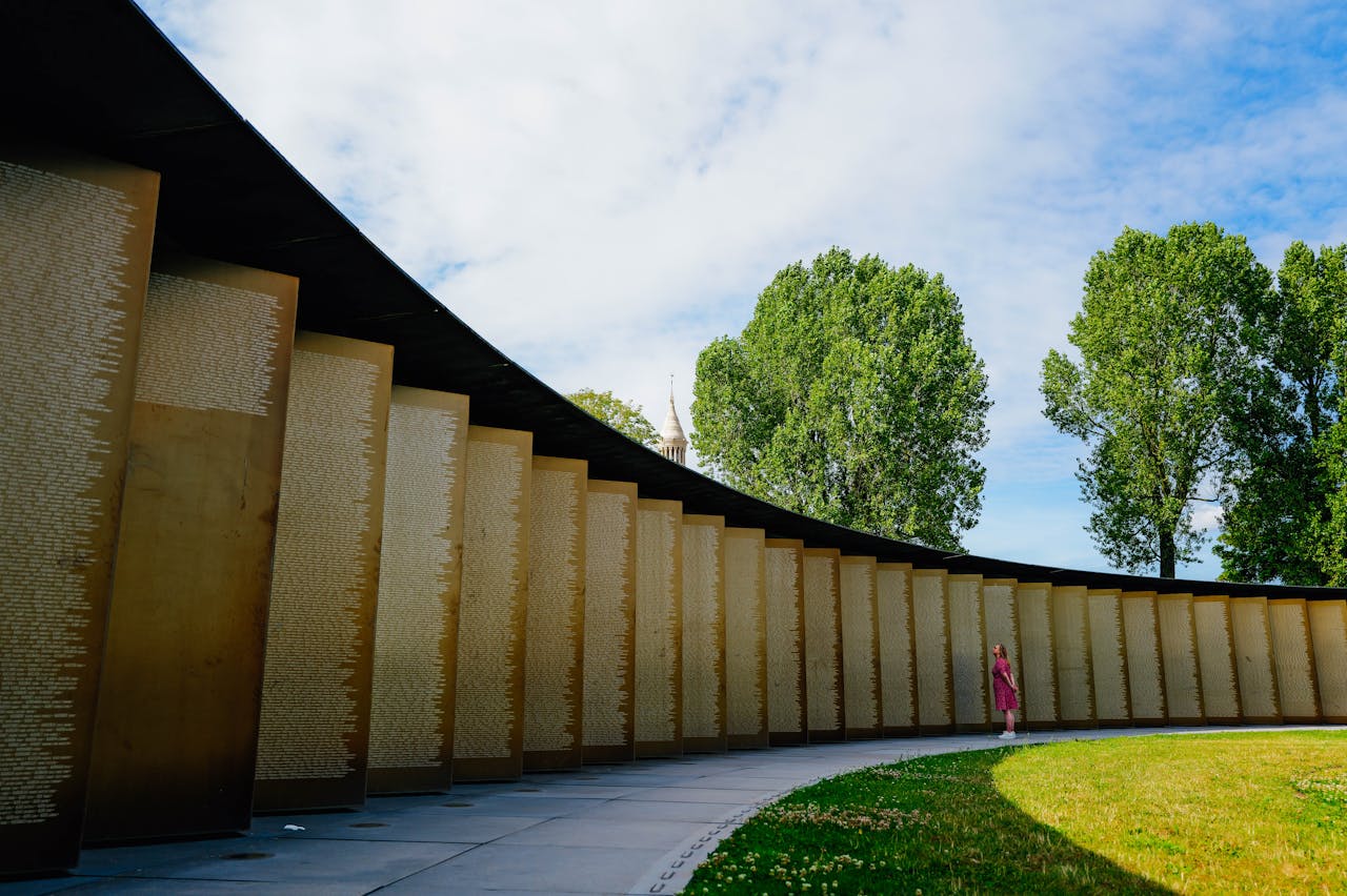A solitary figure in red stands near a modern curved monument wall on a sunny day with trees.